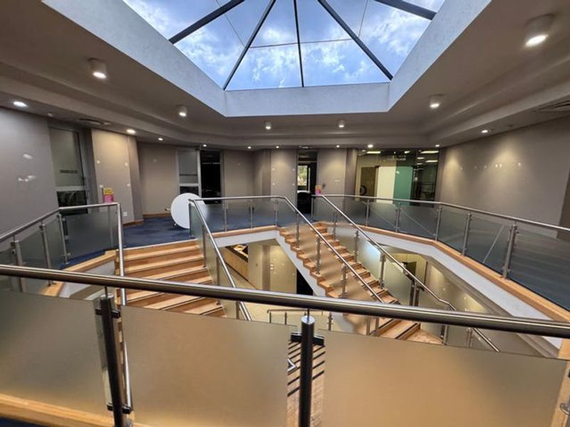 Spacious industrial building atrium featuring a central staircase, glass balustrades, and a large overhead skylight providing natural illumination.