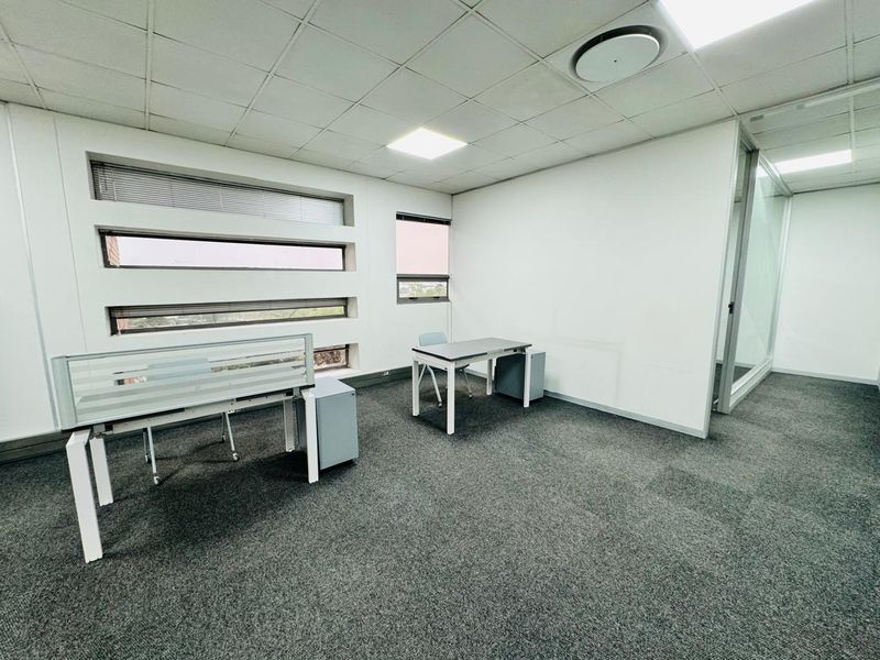 A bright, open-plan office space featuring carpeted flooring, a suspended ceiling with recessed lighting, and glass partition walls.