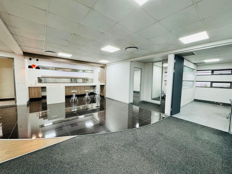 Modern open-plan office space featuring a built-in kitchenette, glass partitions, recessed lighting, and a mix of polished tile and carpet flooring.