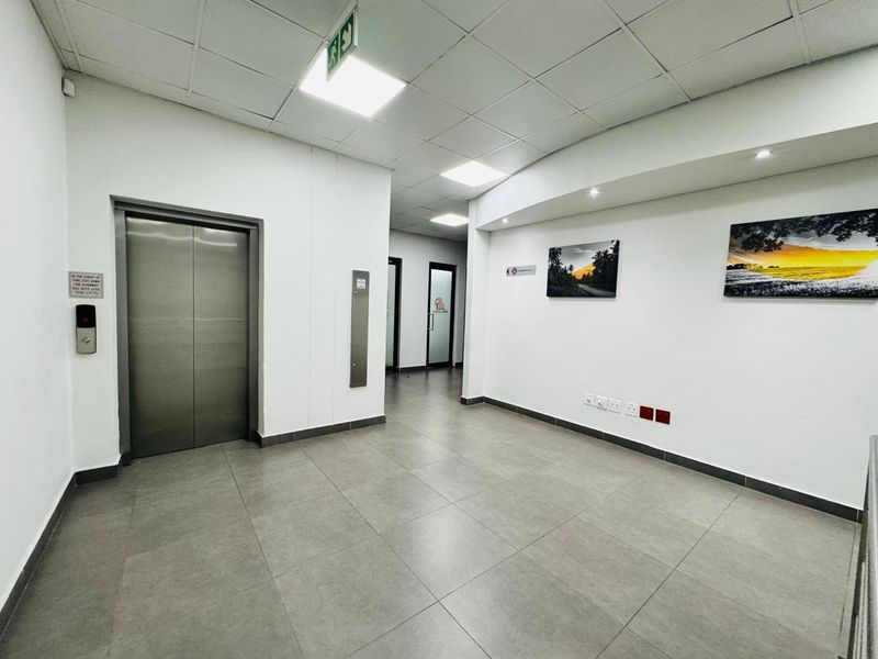 A clean, modern office building hallway featuring tiled flooring, a stainless steel elevator, and recessed lighting in a suspended ceiling.