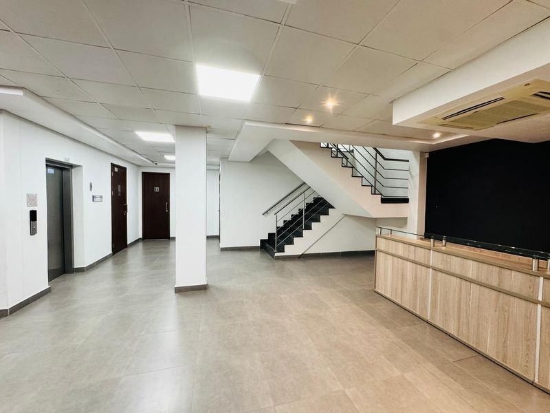 A modern, spacious office lobby featuring a built-in reception desk, tiled flooring, recessed lighting, and a central staircase leading to the upper level.