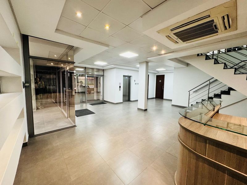 Modern office building entrance lobby featuring a reception desk, elevator access, staircase, and glass-enclosed entry area with neutral tiled flooring.