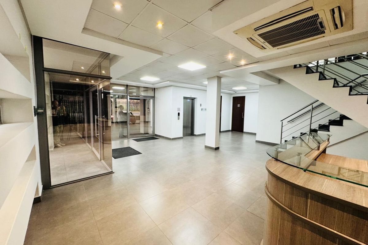 Modern office building entrance lobby featuring a reception desk, elevator access, staircase, and glass-enclosed entry area with neutral tiled flooring.
