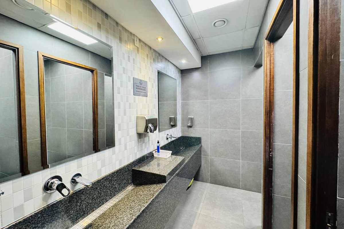 A modern office washroom featuring a long granite vanity with wall-mounted faucets, tiled walls, and recessed lighting in a drop ceiling.