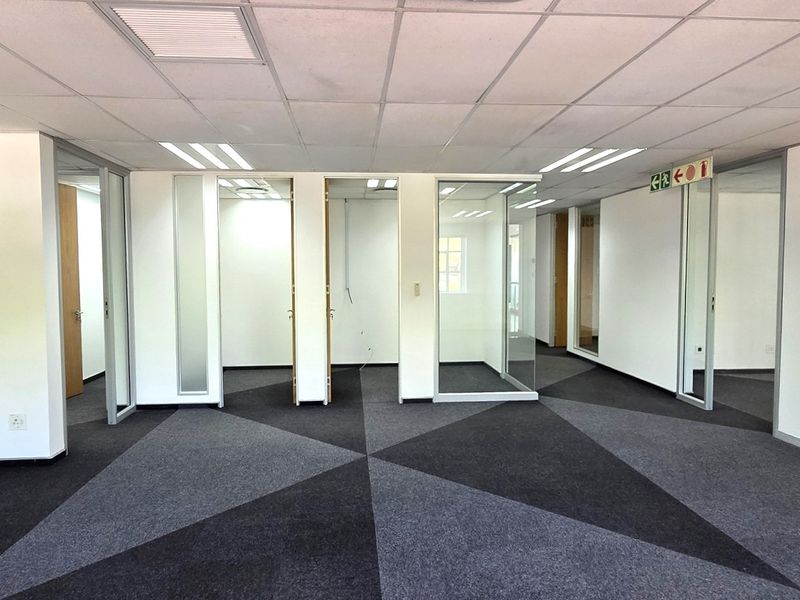 Open-plan office space featuring modern glass partitions, suspended ceilings with recessed lighting, and geometric patterned carpeting.