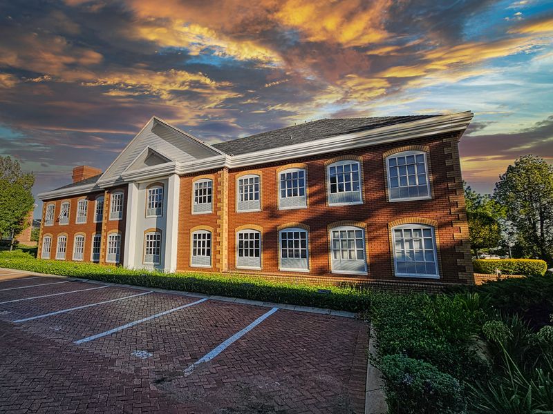 A professional exterior view of a classic brick office building featuring a prominent pedimented entrance and a paved parking area with landscaped surroundings.