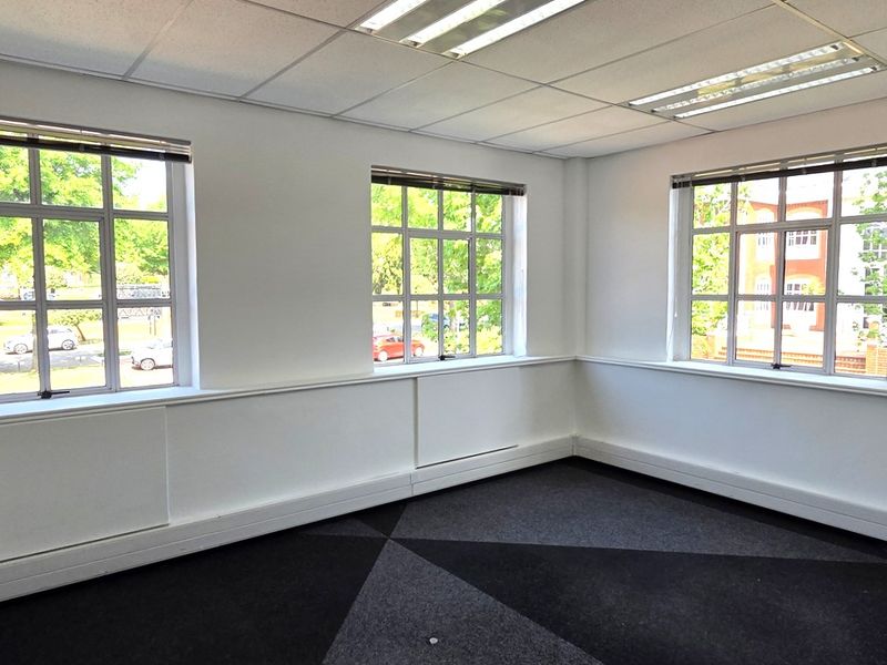 A bright, empty office space featuring large multi-pane windows, a suspended ceiling with recessed lighting, and patterned carpet tiles.