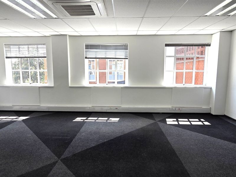 A bright, empty office space featuring a suspended ceiling with recessed lighting, ducted air conditioning, patterned carpet tiles, and large windows with venetian blinds.