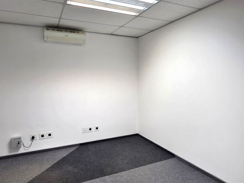 A clean, empty office space featuring carpeted flooring, a suspended ceiling with recessed lighting, and a wall-mounted air conditioning unit.