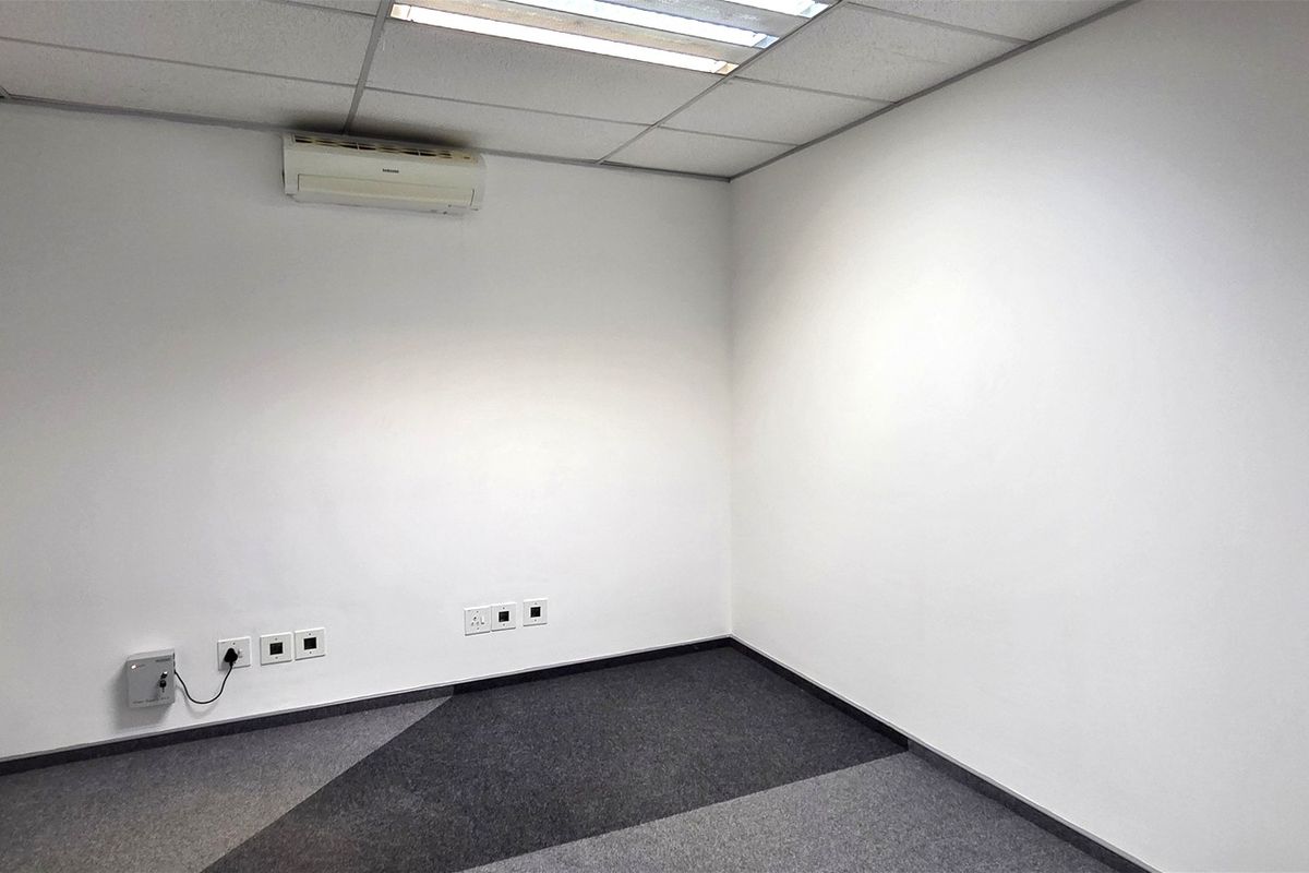 A clean, empty office space featuring carpeted flooring, a suspended ceiling with recessed lighting, and a wall-mounted air conditioning unit.