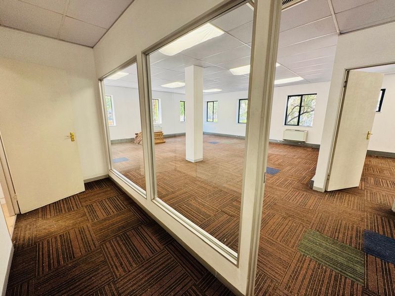 An open-plan office space featuring glass partitions, suspended ceilings with recessed lighting, and carpet tile flooring.