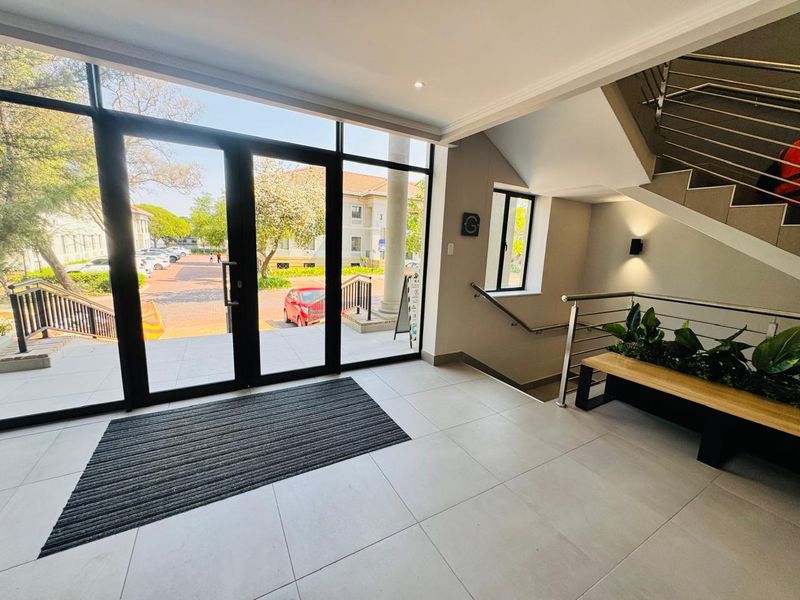 Bright and modern office building entrance featuring large glass doors, tiled flooring, and a contemporary staircase with stainless steel railings.