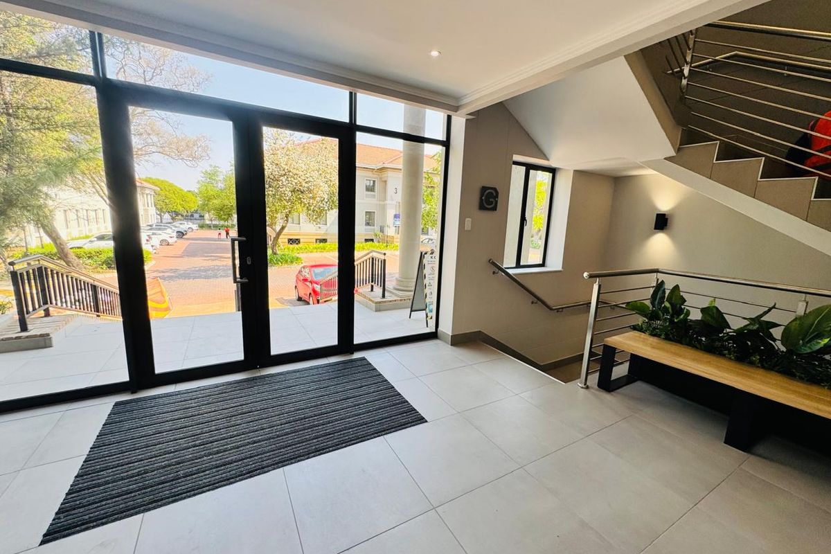 Bright and modern office building entrance featuring large glass doors, tiled flooring, and a contemporary staircase with stainless steel railings.