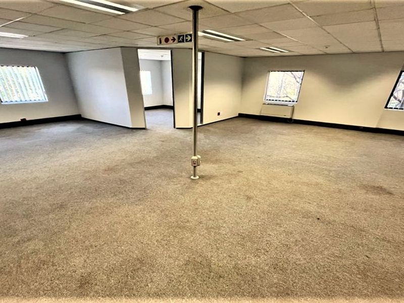 An empty, open-plan office space featuring a suspended ceiling, recessed lighting, and carpeted flooring with a central support pole.