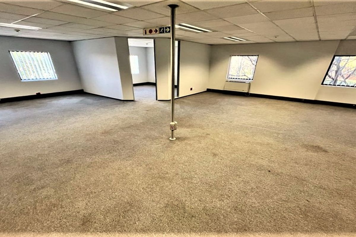 An empty, open-plan office space featuring a suspended ceiling, recessed lighting, and carpeted flooring with a central support pole.