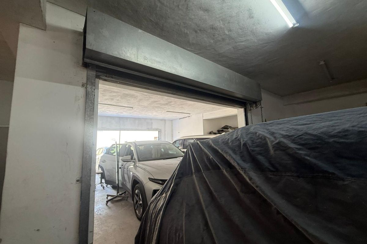 Industrial garage space featuring a metal roller shutter door, concrete flooring, and fluorescent lighting, suitable for vehicle storage or workshop use.