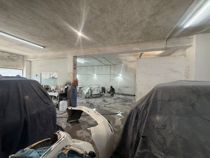 Spacious industrial workshop area featuring concrete flooring, exposed structural beams, and surface-mounted lighting, currently utilized for automotive repair and spray painting.