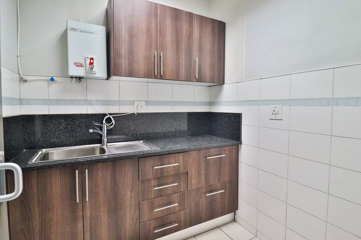 A clean, functional office kitchenette featuring wood-finish cabinetry, granite countertops, a stainless steel sink, and a wall-mounted boiling water unit.