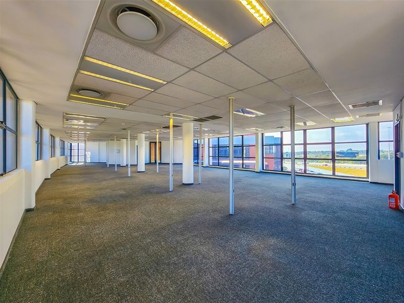 Spacious open-plan office area featuring large windows, carpeted flooring, and a suspended ceiling with integrated lighting and air conditioning.