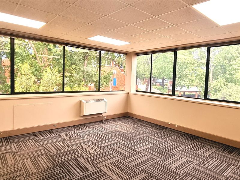 A bright, empty office space featuring large windows with natural views, carpet tile flooring, and a suspended ceiling with recessed lighting.
