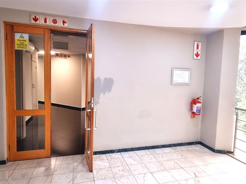 A bright office hallway featuring tiled flooring, a timber-framed entrance door, and essential fire safety equipment with views of surrounding greenery.