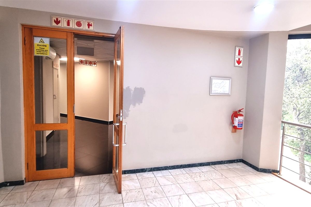 A bright office hallway featuring tiled flooring, a timber-framed entrance door, and essential fire safety equipment with views of surrounding greenery.