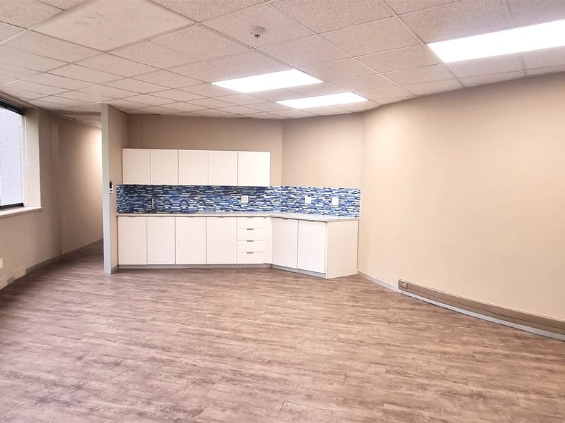 A bright, open-plan office space featuring a built-in kitchenette with tiled backsplash, suspended ceiling, and wood-look laminate flooring.