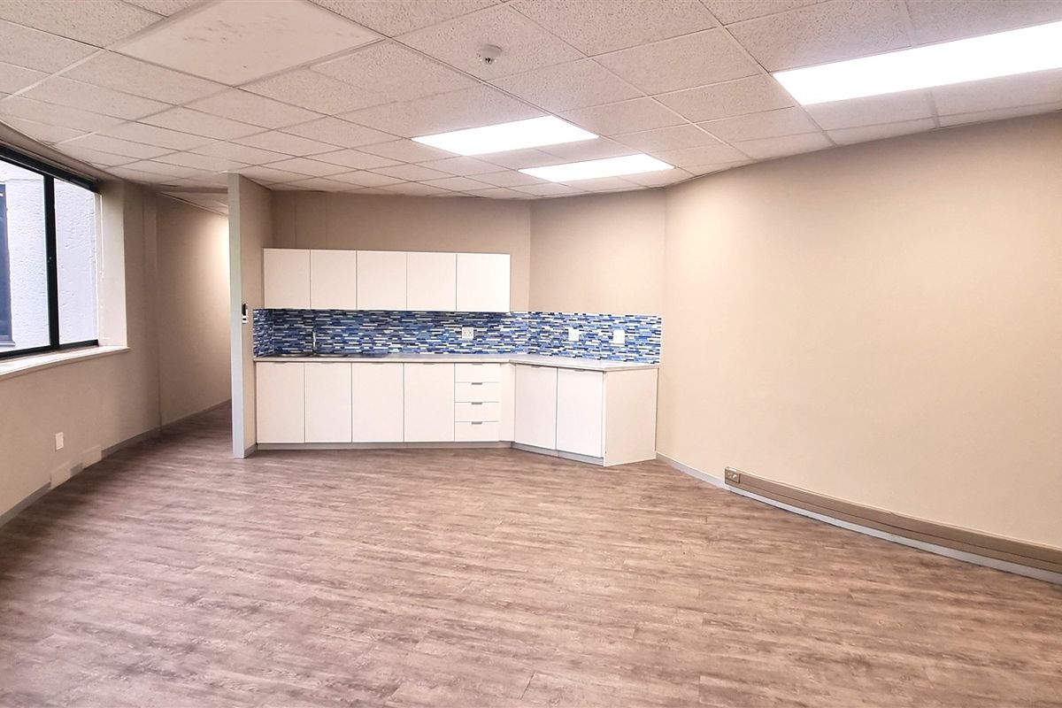 A bright, open-plan office space featuring a built-in kitchenette with tiled backsplash, suspended ceiling, and wood-look laminate flooring.
