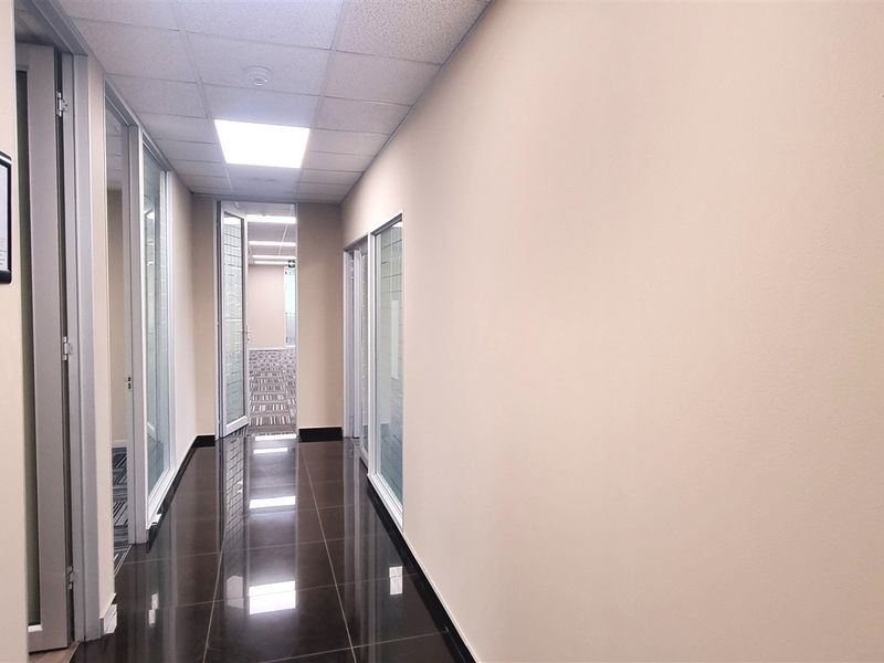 A clean, modern office hallway featuring polished dark floor tiles, suspended ceiling panels, and glass-partitioned office entrances.