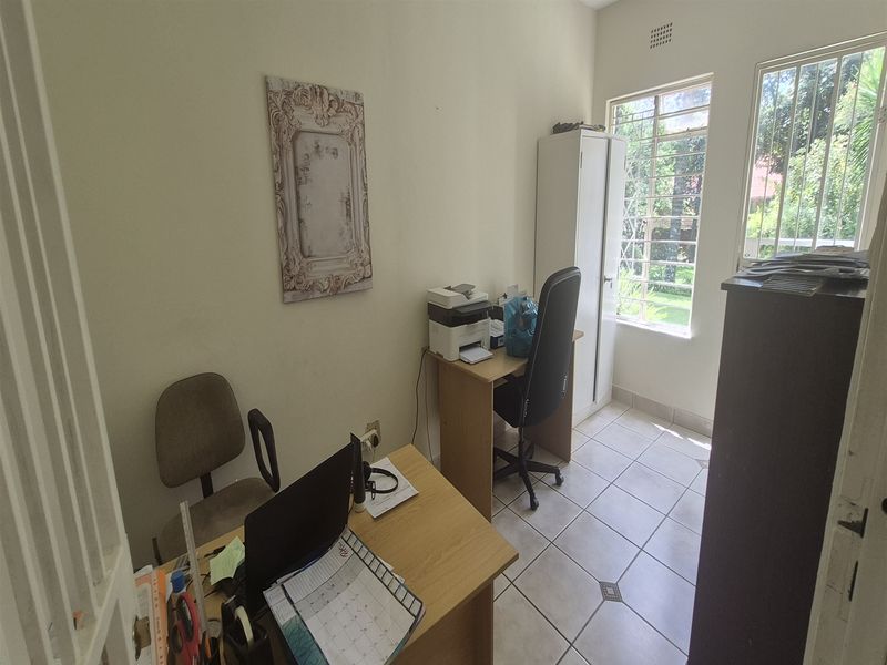 A functional office space featuring tiled flooring and large windows with security bars, providing natural light and a view of the garden.