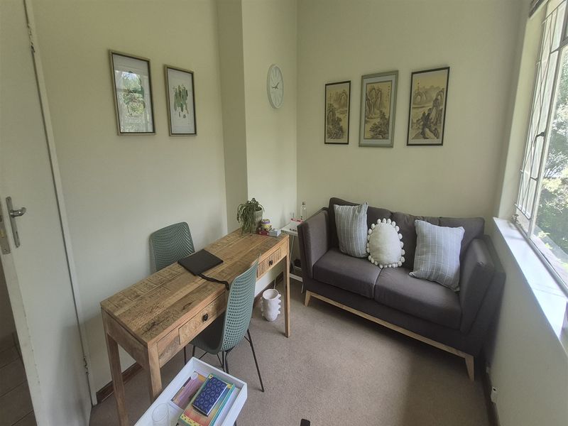 A compact office space featuring neutral walls, carpeted flooring, and a large window providing natural light.