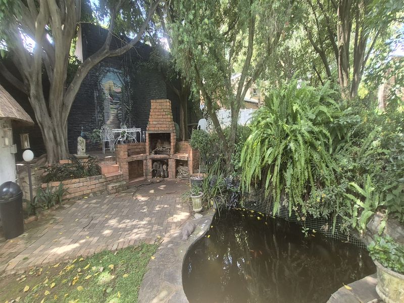A lush, shaded garden area featuring a brick built-in braai, paved patio space, and an ornamental pond surrounded by mature trees and greenery.