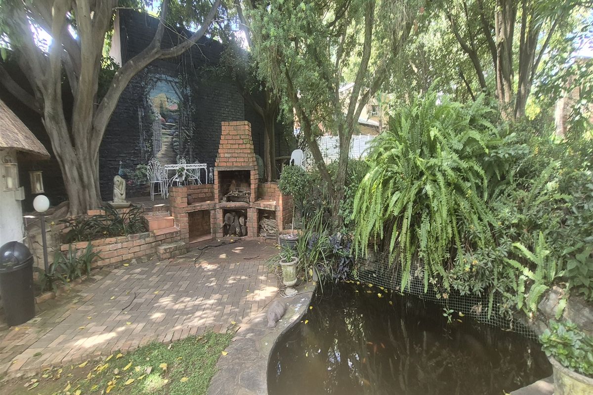 A lush, shaded garden area featuring a brick built-in braai, paved patio space, and an ornamental pond surrounded by mature trees and greenery.