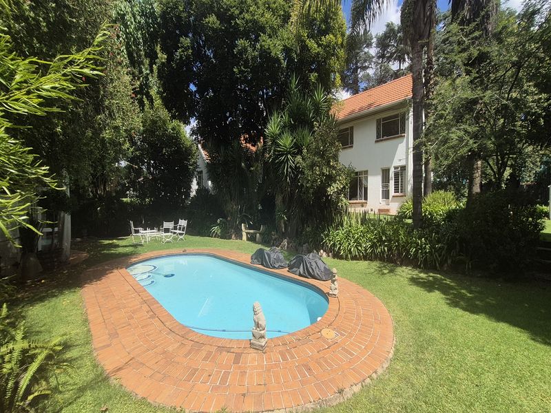 A well-maintained outdoor pool area featuring a classic oval swimming pool with brick coping, surrounded by a lush, established garden and mature trees.
