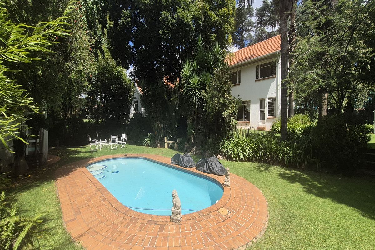 A well-maintained outdoor pool area featuring a classic oval swimming pool with brick coping, surrounded by a lush, established garden and mature trees.