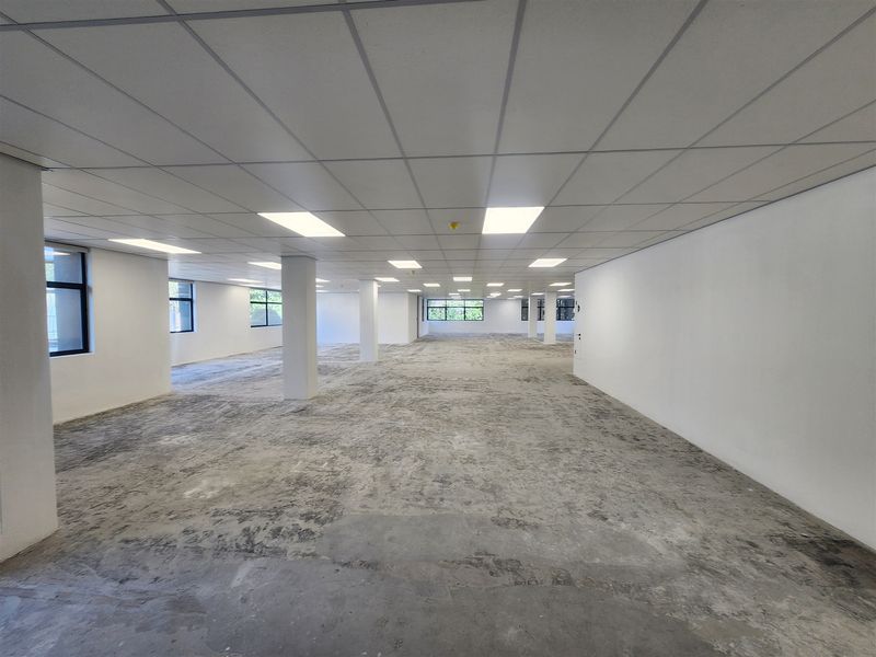 A large, open-plan office space featuring a suspended ceiling with recessed lighting, structural columns, and large windows, currently awaiting final flooring installation.