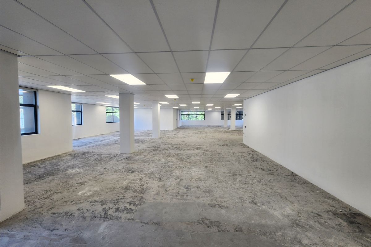 A large, open-plan office space featuring a suspended ceiling with recessed lighting, structural columns, and large windows, currently awaiting final flooring installation.