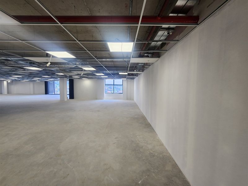 A large, open-plan office space in shell condition featuring exposed concrete ceilings, suspended grid framework, and polished concrete flooring with natural light.