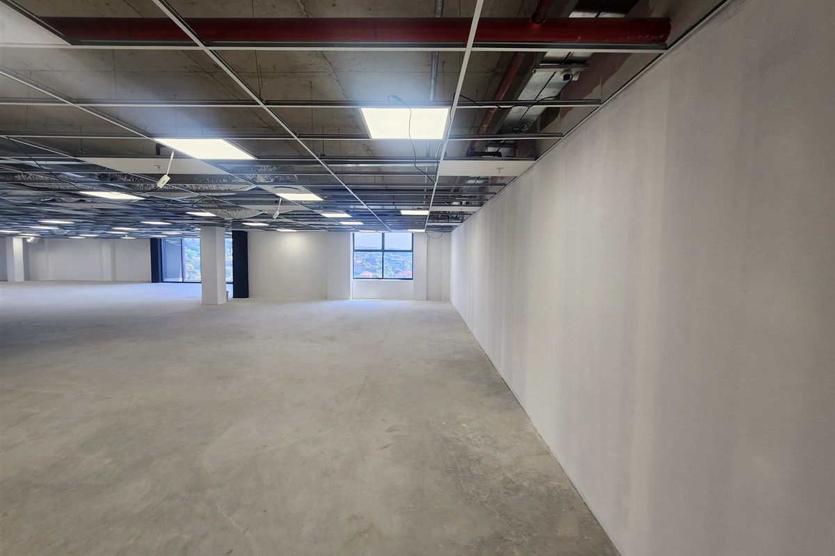 A large, open-plan office space in shell condition featuring exposed concrete ceilings, suspended grid framework, and polished concrete flooring with natural light.
