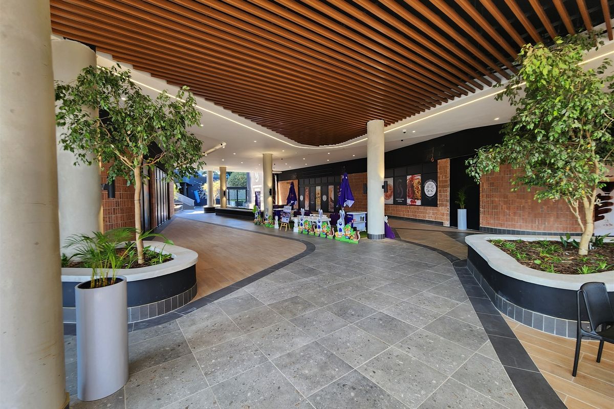 Modern office building entrance lobby featuring a slatted timber ceiling, concrete columns, and integrated planters with a mix of stone and wood-look flooring.