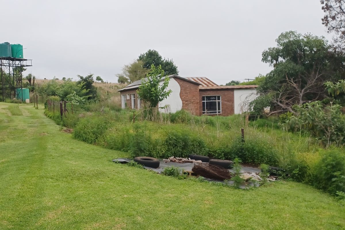 Rear view of a farm property featuring a brick building, corrugated metal roof, and elevated water storage tanks set within an overgrown garden area.