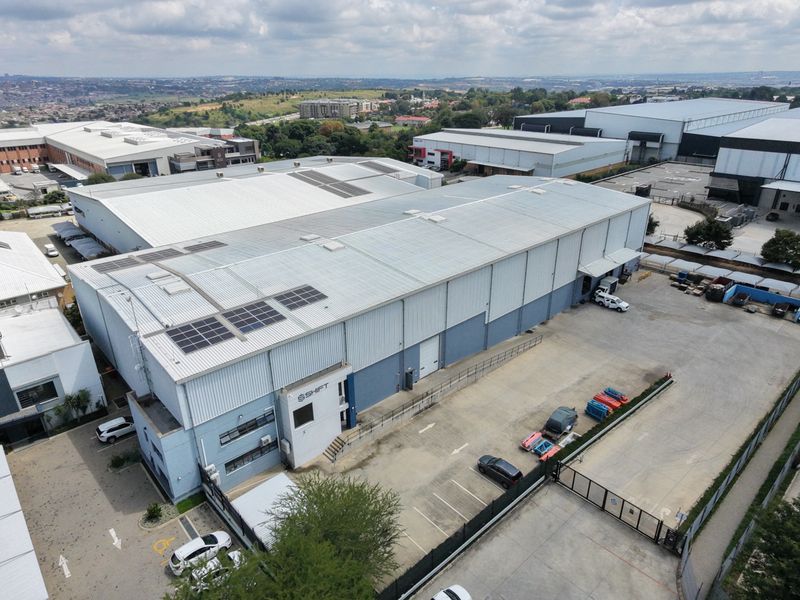 Aerial view of an industrial warehouse facility featuring a metal roof with solar panels, paved parking, and secure perimeter fencing.