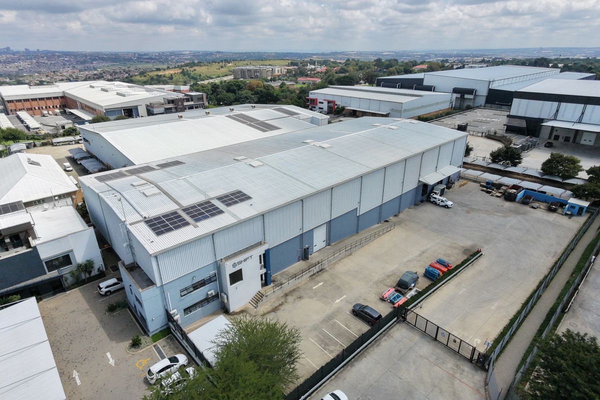 Aerial view of an industrial warehouse facility featuring a metal roof with solar panels, paved parking, and secure perimeter fencing.