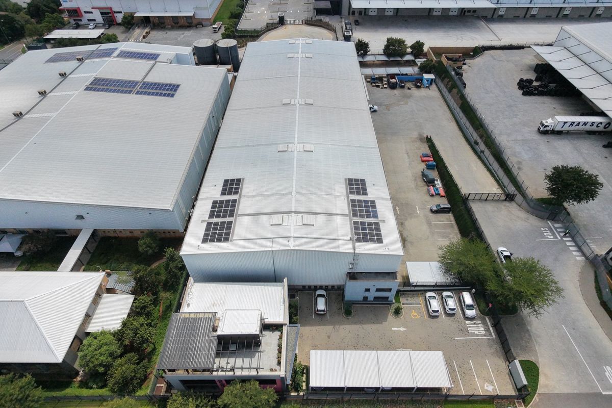 Aerial view of an industrial property featuring a large warehouse with solar panels, paved parking area, and covered parking bays.
