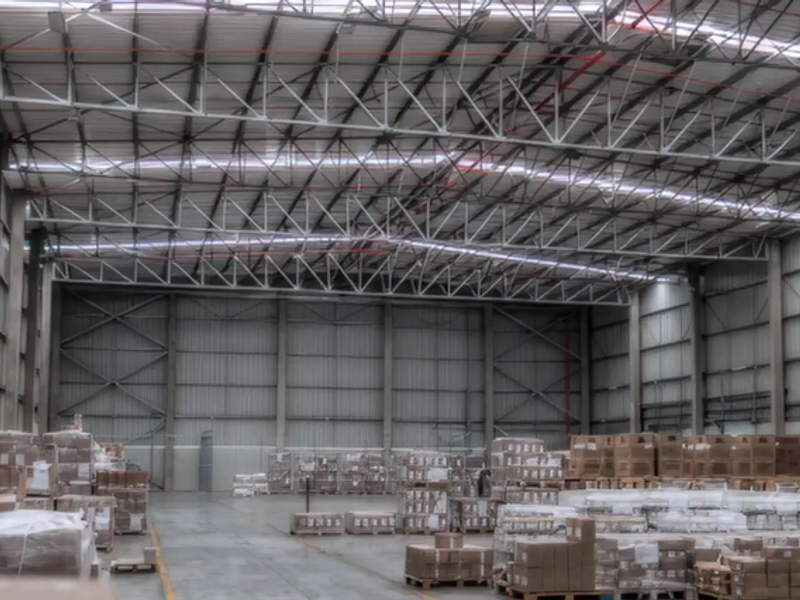 Spacious industrial warehouse featuring high ceilings with exposed steel trusses, corrugated metal walls, and durable concrete flooring, suitable for storage or manufacturing operations.