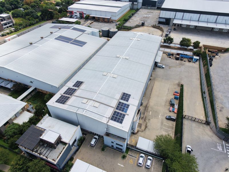 Aerial view of an industrial property featuring large warehouse buildings with solar panel installations, paved parking areas, and secure perimeter fencing.