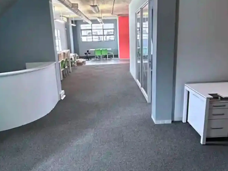Open plan office space featuring exposed industrial ceiling ducting, carpeted flooring, and a modern glass partition wall.