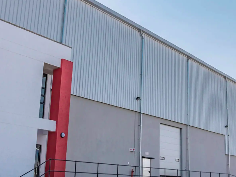 Exterior view of an industrial warehouse facility featuring corrugated metal cladding, a roller shutter door, and a modern office entrance section.