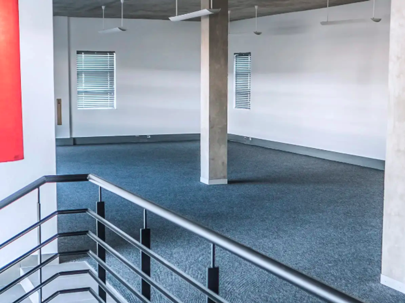 Spacious open-plan industrial office area featuring exposed concrete ceilings, structural columns, carpeted flooring, and large windows with venetian blinds.