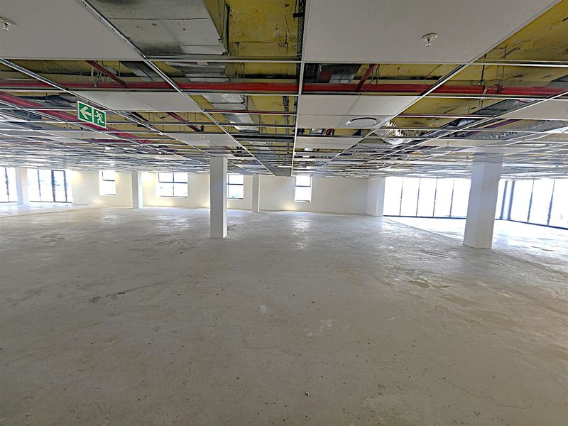 Expansive open-plan office space featuring an unfinished ceiling with exposed services, structural columns, and large windows providing ample natural light.